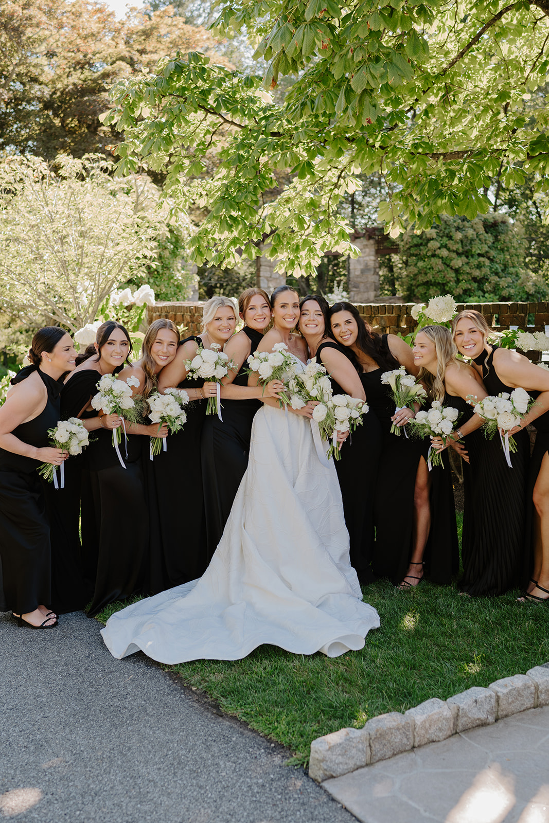 Bride and bridesmaids in black gowns smiling happily while holding white rose bouquets in the garden.